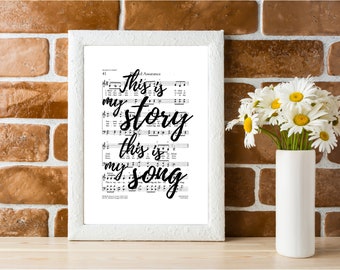 May include: A framed print with the text "This is my story, this is my song" on a white background. The print is framed in a white frame and is sitting on a wooden surface. There are white flowers in a white vase to the right of the print.