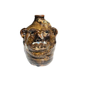 May include: A brown ceramic jug with a face sculpted on the front. The face has a brown beard, a brown mustache, and a brown nose. The jug has a brown glaze with white and blue accents.