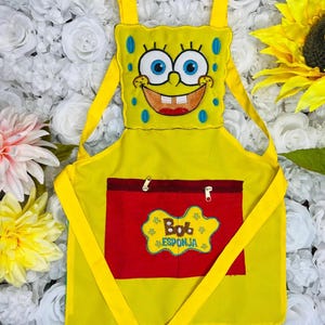 May include: Yellow children's apron with a red pocket and an embroidered image of Spongebob Squarepants. The pocket has two zippers and the text "Bob Esponja" embroidered on it.