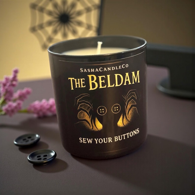 May include: A black candle with the text "The Beldam" in gold lettering, featuring a design of hands and buttons. The candle is from SashaCandleCo and has the phrase "Sew Your Buttons" below the design. Two black buttons are in the foreground.