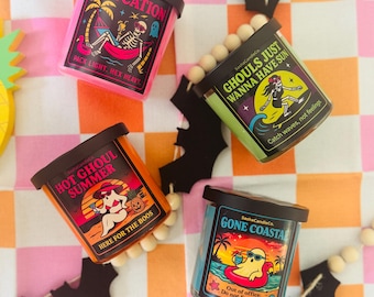 May include: Four scented candles with black lids and colorful labels. The labels feature skeleton and ghost graphics with phrases like "Slaycation" and "Gone Coastal." The candles are arranged on a checkered orange and pink surface.