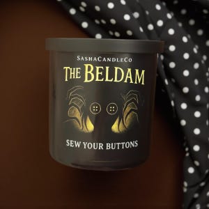 May include: A black candle with a lid, featuring gold text that reads "The Beldam" and "Sew Your Buttons." The candle has an illustration of hands and buttons. The brand name "SashaCandleCo" is also visible.