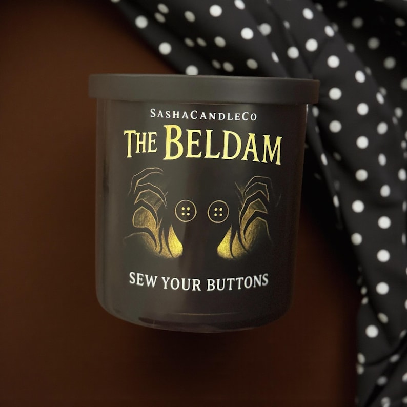 May include: A black candle with a lid, featuring gold text that reads "The Beldam" and "Sew Your Buttons." The candle has an illustration of hands and buttons. The brand name "SashaCandleCo" is also visible.