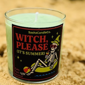 May include: A green candle in a clear glass jar with a black label that reads "Witch, Please (It's Summer)". The label features a skeleton in sunglasses and a witch hat, relaxing on a floatie. The candle is on a sandy surface.