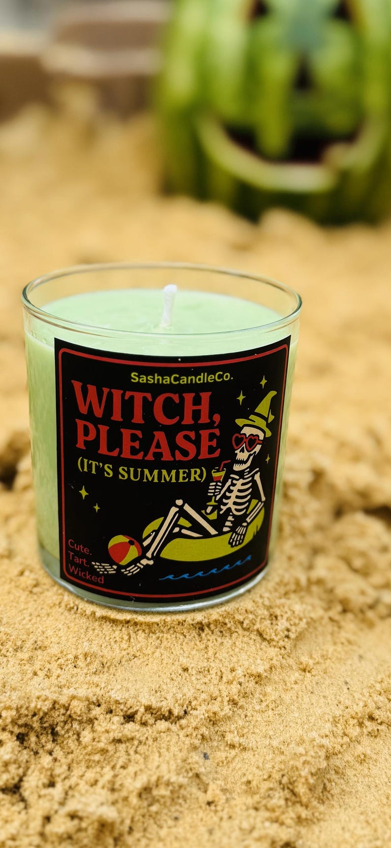 May include: A green candle in a clear glass jar with a black label that reads "Witch, Please (It's Summer)". The label features a skeleton in sunglasses and a witch hat, relaxing on a floatie. The candle is on a sandy surface.