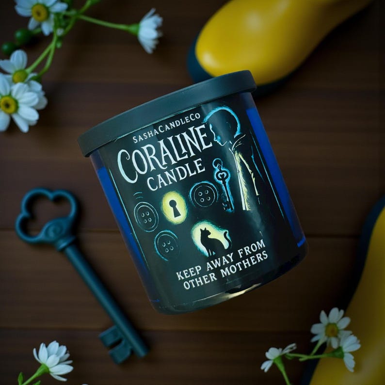 May include: A cylindrical candle with a black lid and a blue label featuring the text "Coraline Candle" and illustrations of buttons, a key, and a cat. The candle is surrounded by a decorative key, daisies, and yellow boots.
