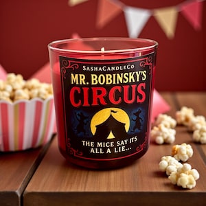 May include: A red candle in a glass jar with the text "MR. BOBINSKY'S CIRCUS" on the front. The candle is next to a bowl of popcorn and some loose popcorn kernels. The background is red.