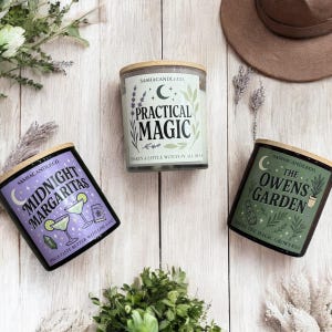 May include: Three scented candles with wooden lids are displayed on a light wood surface. The labels feature the text "Midnight Margaritas", "Practical Magic", and "The Owens Garden". Each candle has a unique design with illustrations and text.