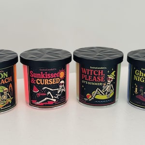 May include: Four scented candles with black lids and labels featuring Halloween-themed graphics. The candles have different colors, including orange, pink, green, and blue. Each candle has a unique design with text like "Hex on the Beach" and "Ghoul's Night In."