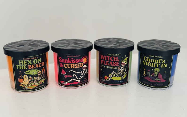 May include: Four scented candles with black lids and labels featuring Halloween-themed graphics. The candles have different colors, including orange, pink, green, and blue. Each candle has a unique design with text like "Hex on the Beach" and "Ghoul's Night In."