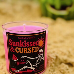 May include: A pink candle in a clear glass jar with a black label that reads "Sunkissed & Cursed." The label features a skeleton wearing sunglasses and a watermelon slice. The candle is on sand, with a blurred watermelon in the background.
