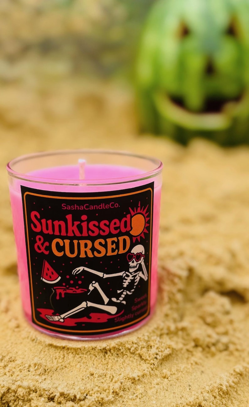 May include: A pink candle in a clear glass jar with a black label that reads "Sunkissed & Cursed." The label features a skeleton wearing sunglasses and a watermelon slice. The candle is on sand, with a blurred watermelon in the background.