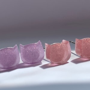May include: A set of four cat-shaped stud earrings. Two are a light purple color with glitter, and two are a light pink color with glitter.