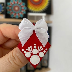 May include: A small, red, diamond-shaped wooden ornament with a white bow and a white painted design. The design features a series of circles and swirls.