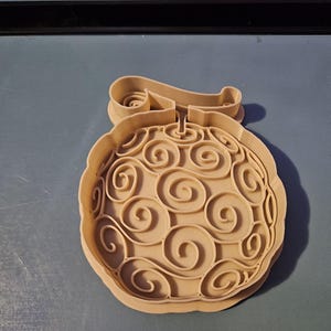 May include: A tan-colored cookie cutter in the shape of a stylized fruit with a swirling pattern. The cutter has a decorative scroll at the top. The design is reminiscent of a cartoon or anime style.