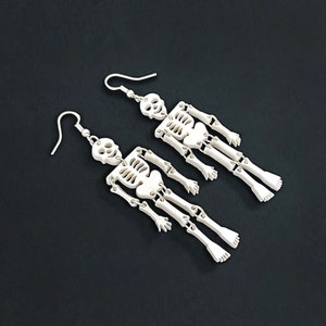 Puede incluir: Un par de pendientes de esqueleto hechos de pl&aacute;stico blanco. Los pendientes son de estilo colgante y presentan un dise&ntilde;o de esqueleto con extremidades articuladas.