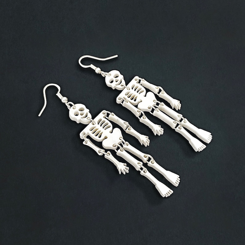 Puede incluir: Un par de pendientes de esqueleto hechos de pl&aacute;stico blanco. Los pendientes son de estilo colgante y presentan un dise&ntilde;o de esqueleto con extremidades articuladas.