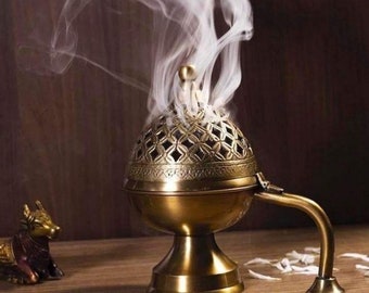 May include: A gold metal incense burner with a decorative, openwork design. The burner has a handle and sits on a base. Smoke is rising from the burner.