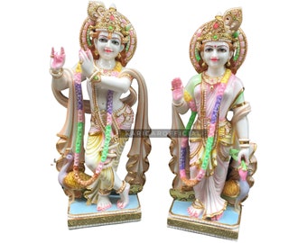 May include: A pair of white marble statues of Hindu deities, Radha and Krishna, standing side-by-side. The statues are intricately carved with colorful details and elaborate headdresses. Krishna is holding a flute and Radha is holding a flower.