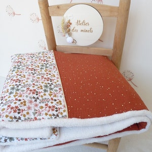 May include: A folded blanket with a floral pattern on one side and a rust-colored fabric with gold speckles on the other. The blanket has a white, fluffy trim. The blanket is on a wooden chair.