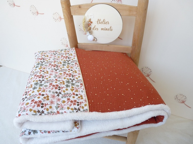 May include: A folded blanket with a floral pattern on one side and a rust-colored fabric with gold speckles on the other. The blanket has a white, fluffy trim. The blanket is on a wooden chair.