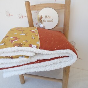 May include: A folded baby blanket with a whimsical fox and forest print in mustard yellow and rust orange. The blanket has a soft, white sherpa lining and is draped over a wooden chair. The blanket features the text "Atelier des minets" on a wooden disc.