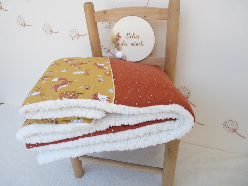May include: A folded baby blanket with a whimsical fox and forest print in mustard yellow and rust orange. The blanket has a soft, white sherpa lining and is draped over a wooden chair. The blanket features the text "Atelier des minets" on a wooden disc.