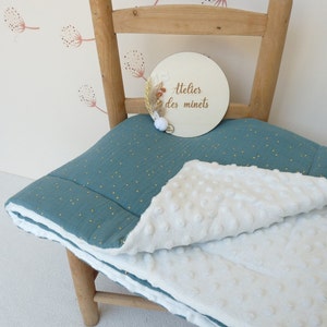 May include: A teal and white baby blanket with a textured, dotted pattern. The blanket is folded over a wooden chair. A wooden sign with the text "atelier des minets" is on the chair.