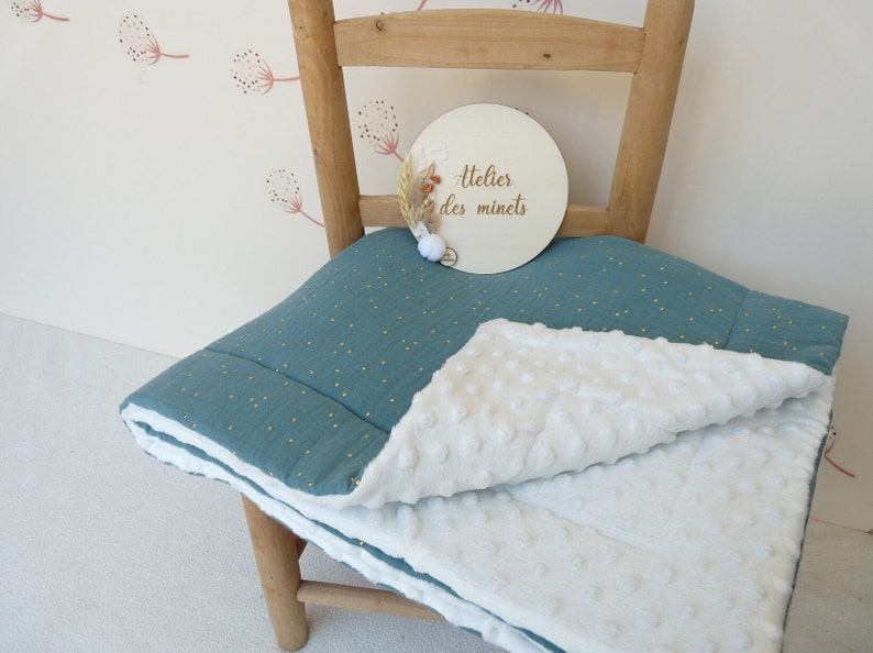 May include: A teal and white baby blanket with a textured, dotted pattern. The blanket is folded over a wooden chair. A wooden sign with the text "atelier des minets" is on the chair.