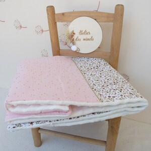 May include: A pink and white baby blanket with gold polka dots and a floral pattern. The blanket is folded over a wooden chair. A wooden sign with the text "Atelier des minets" is attached to the chair.