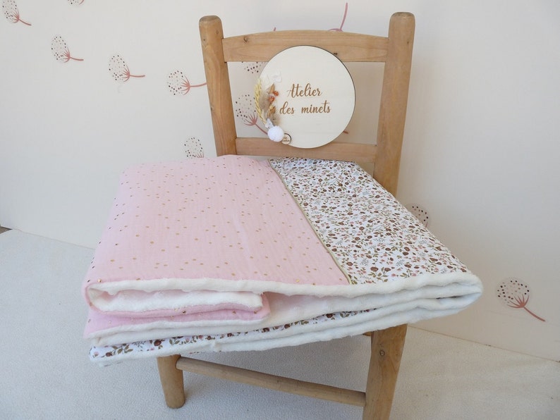 May include: A pink and white baby blanket with gold polka dots and a floral pattern. The blanket is folded over a wooden chair. A wooden sign with the text "Atelier des minets" is attached to the chair.