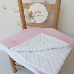 May include: A pink and white baby blanket with gold polka dots is draped over a wooden chair. The blanket has a textured white side and a pink side with gold dots. A wooden sign with the text "Atelier des minets" is attached to the chair.