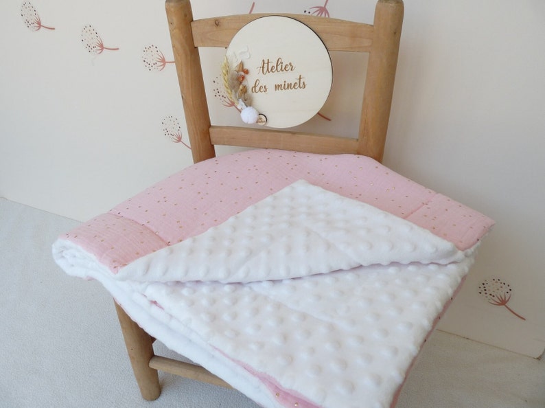 May include: A pink and white baby blanket with gold polka dots is draped over a wooden chair. The blanket has a textured white side and a pink side with gold dots. A wooden sign with the text "Atelier des minets" is attached to the chair.