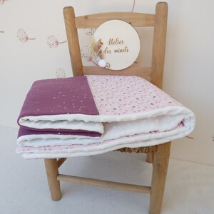 May include: A folded baby blanket with a two-tone design. One half is a deep purple with white star accents, and the other half is light pink with star and heart patterns. The blanket is on a wooden chair.