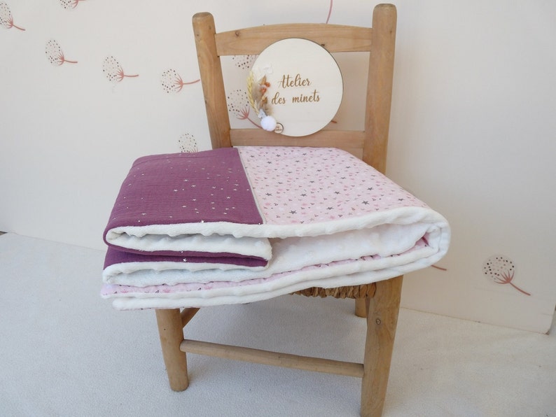 May include: A folded baby blanket with a two-tone design. One half is a deep purple with white star accents, and the other half is light pink with star and heart patterns. The blanket is on a wooden chair.