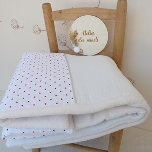 May include: A folded white baby blanket with a red heart pattern on one side and a soft, plush texture on the other. The blanket is layered with a white, textured blanket. A wooden sign with the text "Atelier des minets" is in the background.