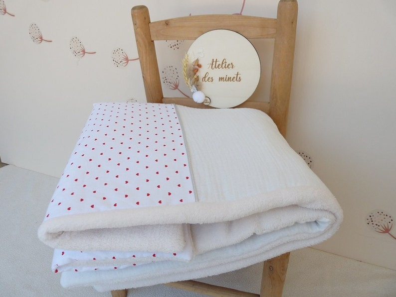 May include: A folded white baby blanket with a red heart pattern on one side and a soft, plush texture on the other. The blanket is layered with a white, textured blanket. A wooden sign with the text "Atelier des minets" is in the background.