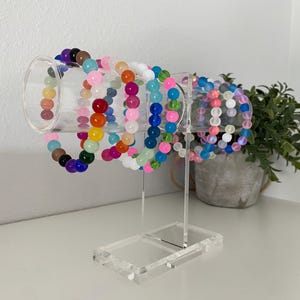 May include: A clear acrylic bracelet display stand holding several beaded bracelets. The bracelets are various colors, including pink, blue, green, and purple. The stand is on a white surface.