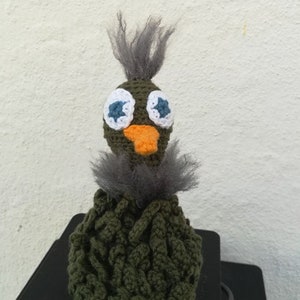 Könnte beinhalten: Ein gehäkeltes Stofftier in Form eines Vogels mit einem flauschigen grauen Kopf, großen weißen Augen mit blauen Pupillen, einem orangefarbenen Schnabel und einem grünen Körper.