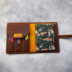 May include: Open brown leather pencil case with a flap closure, holding a set of colored pencils. A notebook with a koi fish pattern is visible inside. The name "Maria" is embossed on a small label.