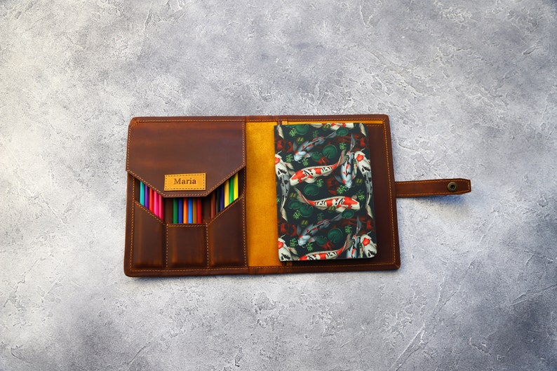 May include: Open brown leather pencil case with a flap closure, holding a set of colored pencils. A notebook with a koi fish pattern is visible inside. The name "Maria" is embossed on a small label.