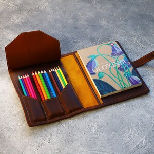 May include: Open brown leather pencil case with a selection of colored pencils and a notebook. The notebook has the word "GLOXINIA" on the cover, along with a floral illustration. The case is open, revealing a yellow interior.