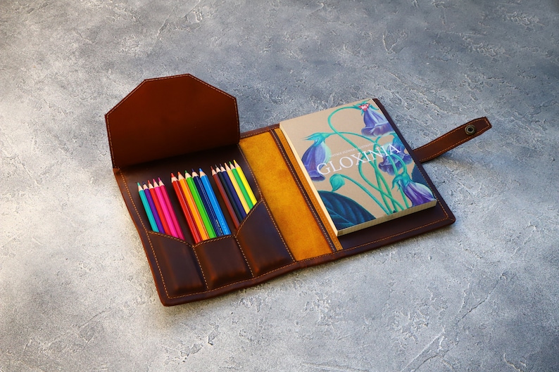 May include: Open brown leather pencil case with a selection of colored pencils and a notebook. The notebook has the word "GLOXINIA" on the cover, along with a floral illustration. The case is open, revealing a yellow interior.