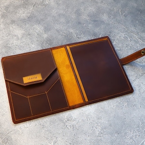 May include: A brown leather portfolio with a flap pocket and a strap closure. The interior features a yellow-orange lining. The name "Maria" is embossed on a small rectangular patch. Orange stitching details the edges and pockets.