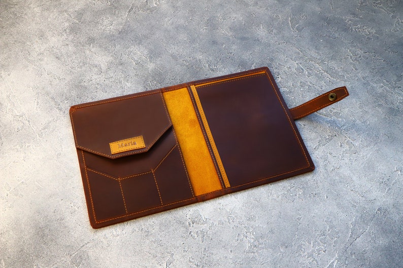 May include: A brown leather portfolio with a flap pocket and a strap closure. The interior features a yellow-orange lining. The name "Maria" is embossed on a small rectangular patch. Orange stitching details the edges and pockets.