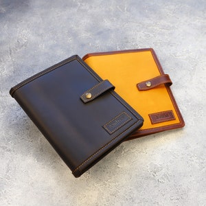May include: Two leather-bound journals. One is dark brown with a stitched border and a brass clasp. The other journal has an orange canvas cover with a brown leather trim and clasp. Both have name tags.