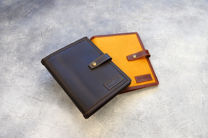 May include: Two leather-bound journals. One is dark brown with a stitched border and a brass clasp. The other journal has an orange canvas cover with a brown leather trim and clasp. Both have name tags.