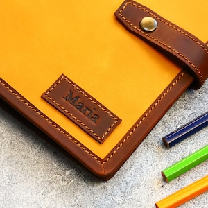 May include: A handcrafted leather journal in a vibrant yellow hue, accented with rich brown trim and a matching strap closure. The journal features the name "Maria" embossed on a small leather patch. Colored pencils are visible.