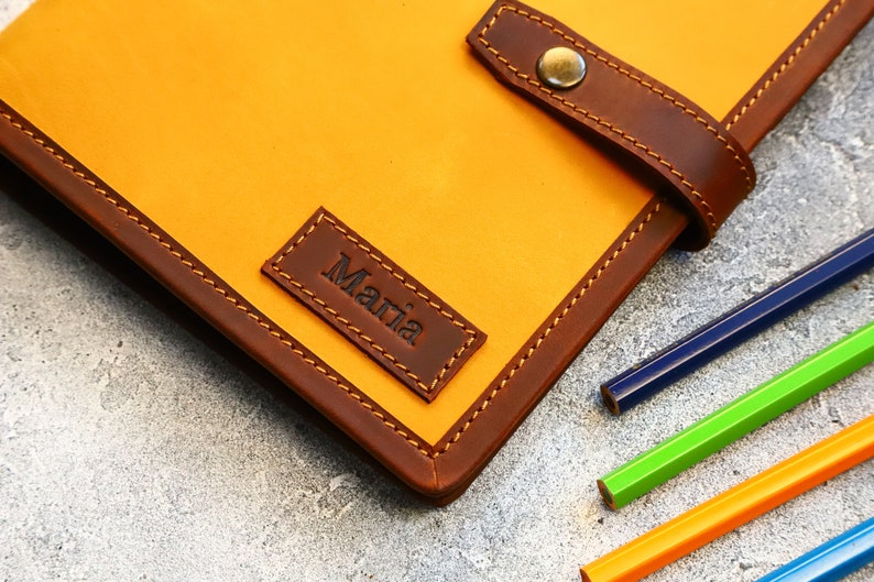 May include: A handcrafted leather journal in a vibrant yellow hue, accented with rich brown trim and a matching strap closure. The journal features the name "Maria" embossed on a small leather patch. Colored pencils are visible.