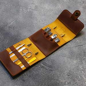 May include: A brown leather travel case with a yellow lining, open to reveal a set of manicure tools, including nail clippers, tweezers, a file, and a cuticle pusher.